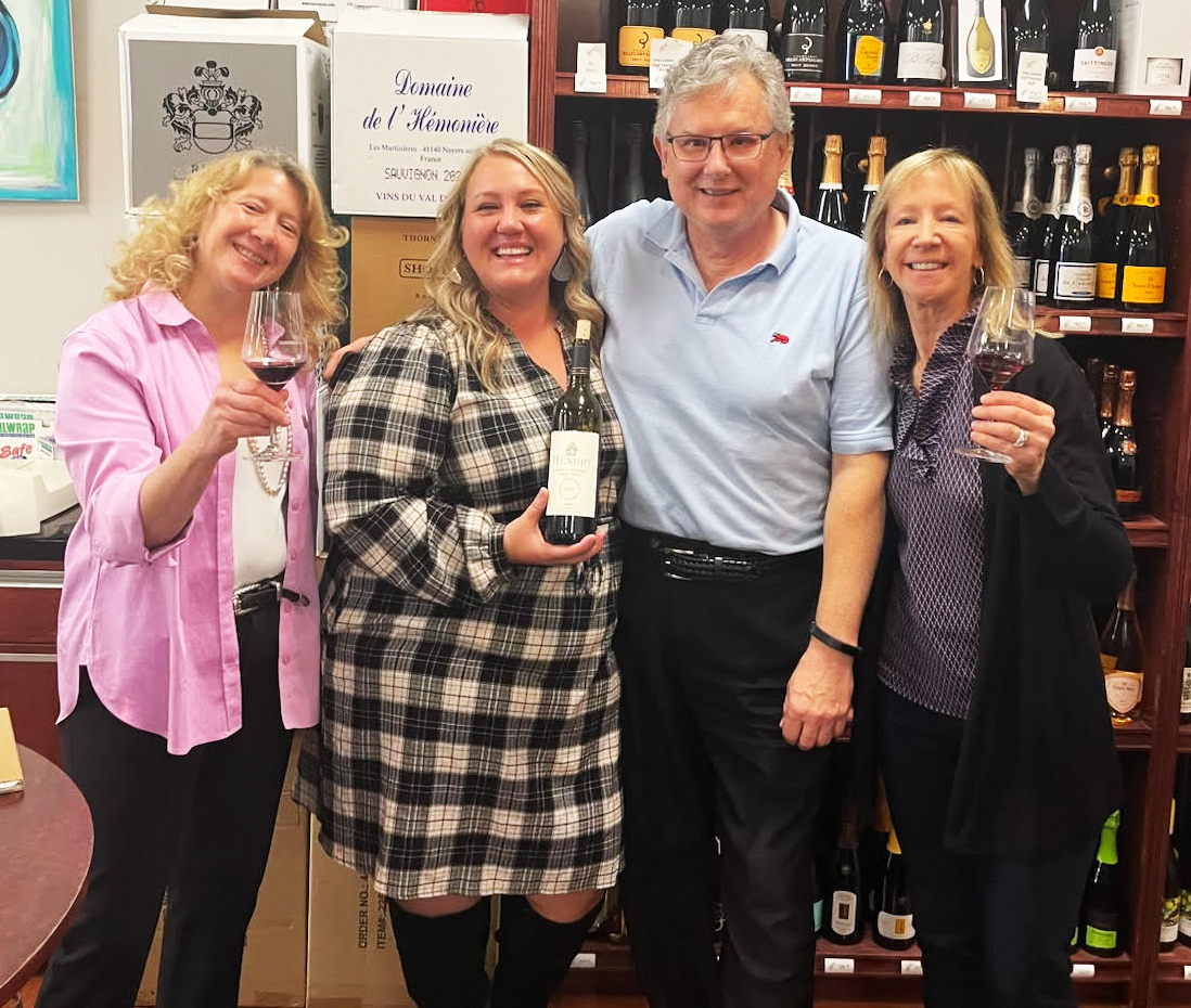 Angela stands with the owners of Virginia's Wine Cabinet with wine bottles behind them